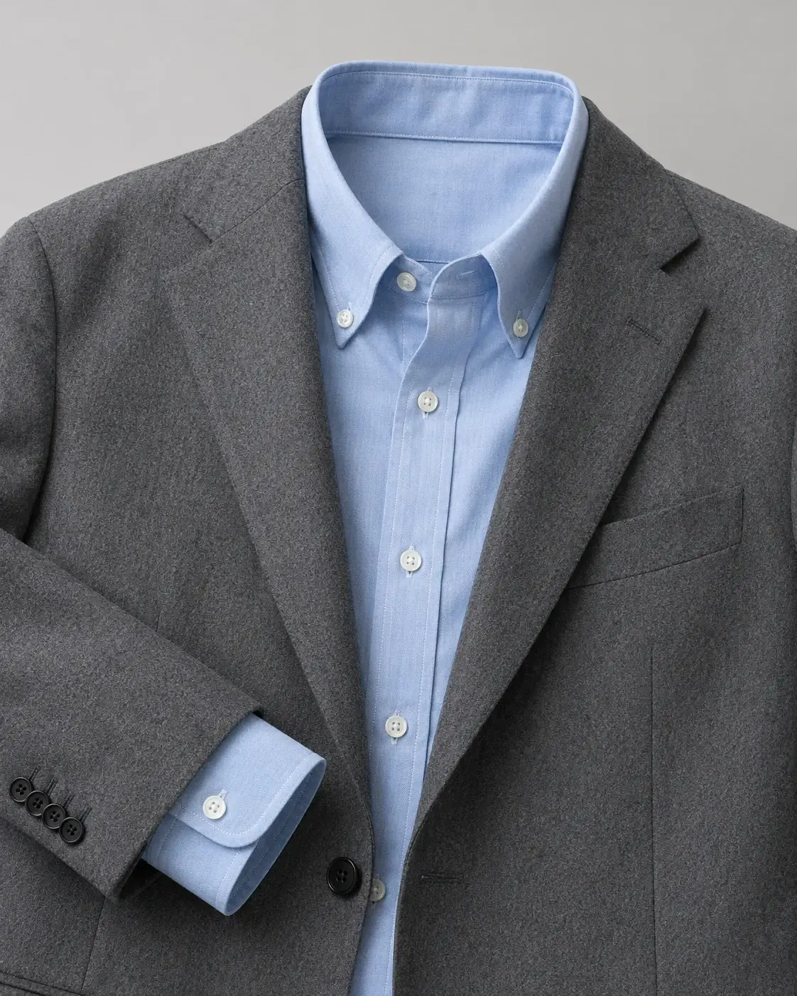 Product shot of a light blue oxford shirt and a grey wool blazer on a plain neutral background, crisp editorial lighting