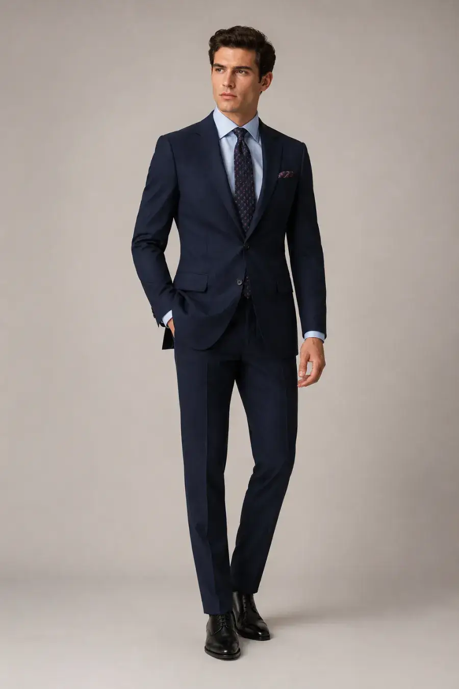 Full body shot of a men's cocktail look — a navy tailored suit with a light blue shirt and patterned tie, on a plain neutral studio background