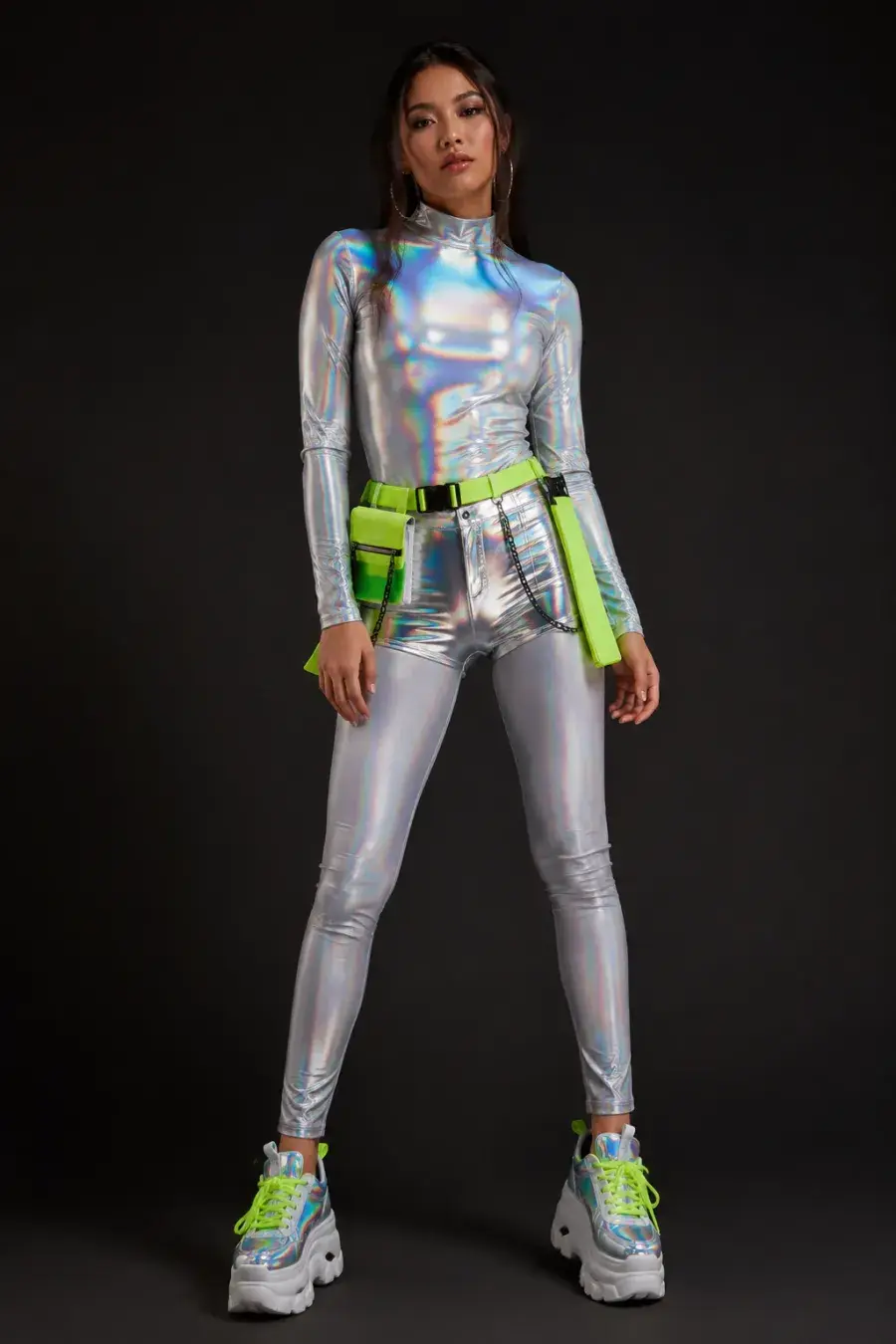 Rave festival outfit: a holographic long-sleeve top, metallic high-waisted shorts, a neon utility belt, and chunky platform sneakers shown on a model against a plain dark studio background, vivid lighting, full body