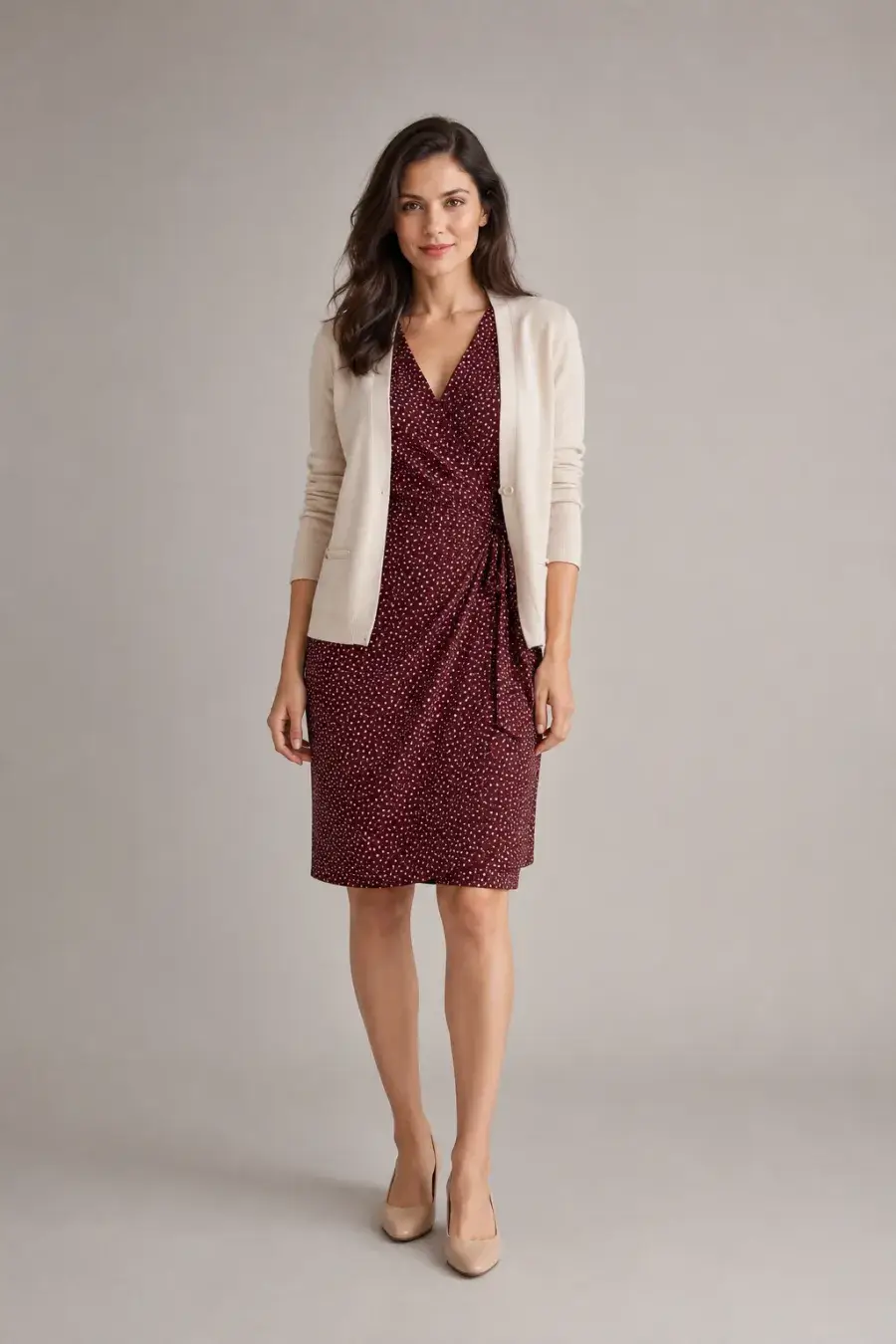 A woman in her late twenties wearing a burgundy knee-length wrap dress with a small white dot print, a cream tailored cardigan, and tan low heels, standing upright facing forward, full body, entire figure from head to feet in frame, centered with headroom